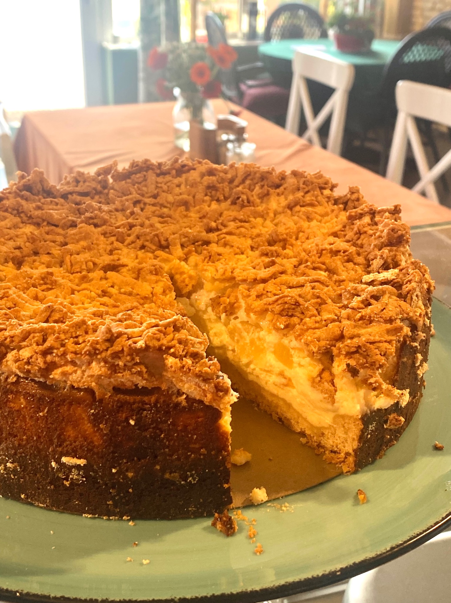 Kawałek wyciętego sernika z kruszonką na zielonym talerzu, w tle stolik z kwiatami. Sernik ma wyraźną warstwę kremową i ciemną podstawę.