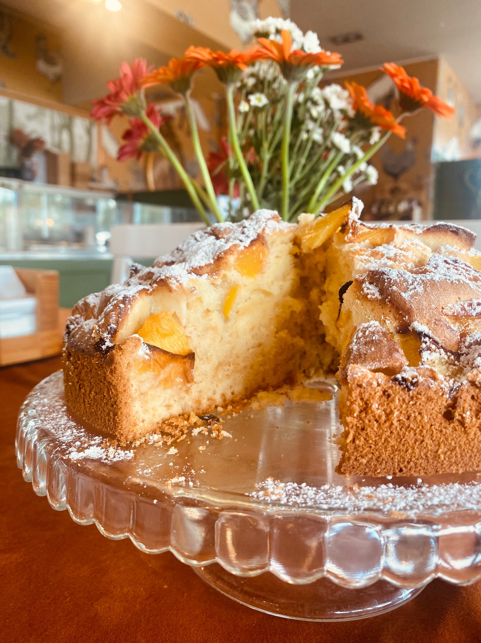 Kawałek ciasta z brzoskwiniami i cukrem pudrem na szklanym stojaku. W tle kwiaty i wyposażenie cukierni.