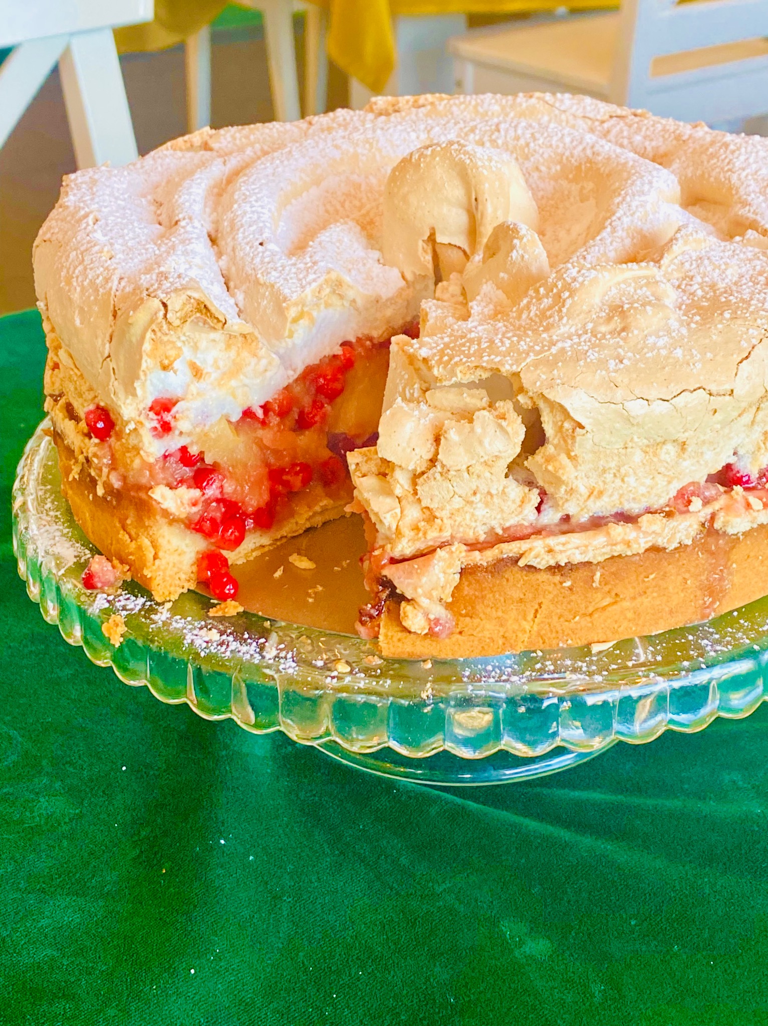 Krojony tort bezowy z owocami na szklanej paterze, posypany cukrem pudrem. Widoczne warstwy ciasta, bezy i czerwonych owoców. Tło stanowią białe krzesła i zielony obrus.