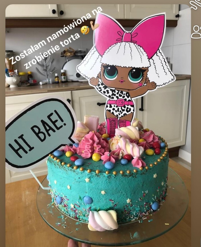 Turkusowy tort z topperem LOL, dekorowany piankami i cukierkami, na szklanej paterze. Napis 'HI BAE!' w dymku.