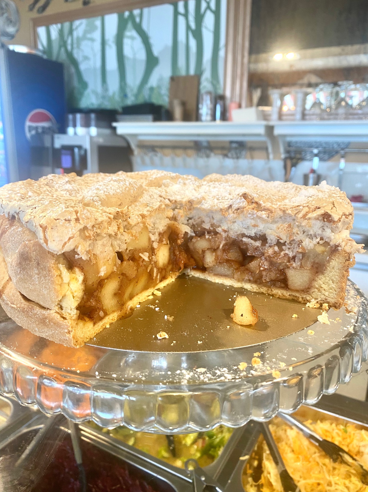 Przekrojone ciasto z jabłkami i bezą na szklanym stojaku, widoczne nadzienie i kruchy spód. W tle elementy wystroju wnętrza, w tym regał z naczyniami.