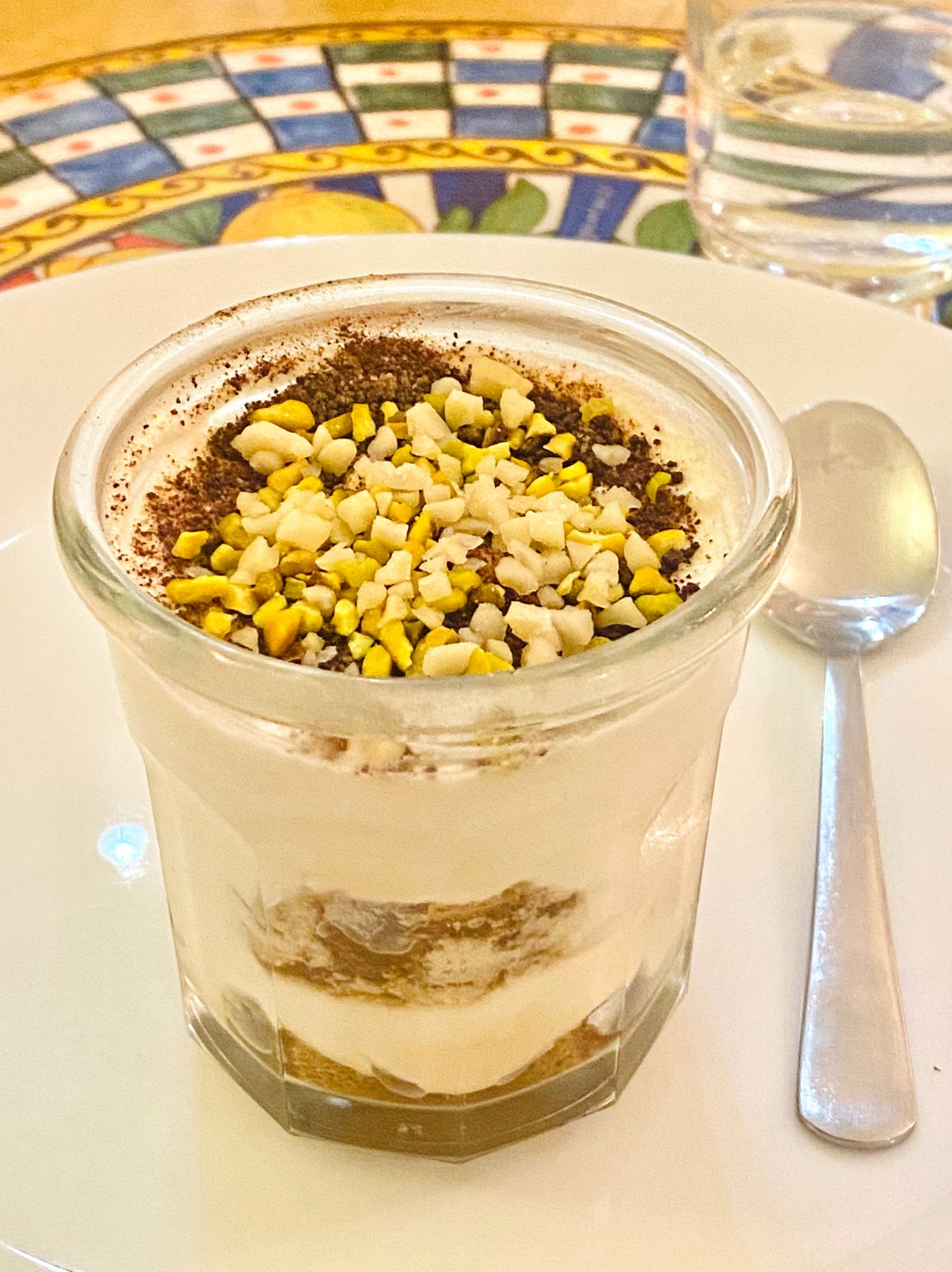Deser tiramisu w szklance, posypany orzechami i kakao, obok łyżeczka. W tle woda w szklance i wzorzysty obrus.