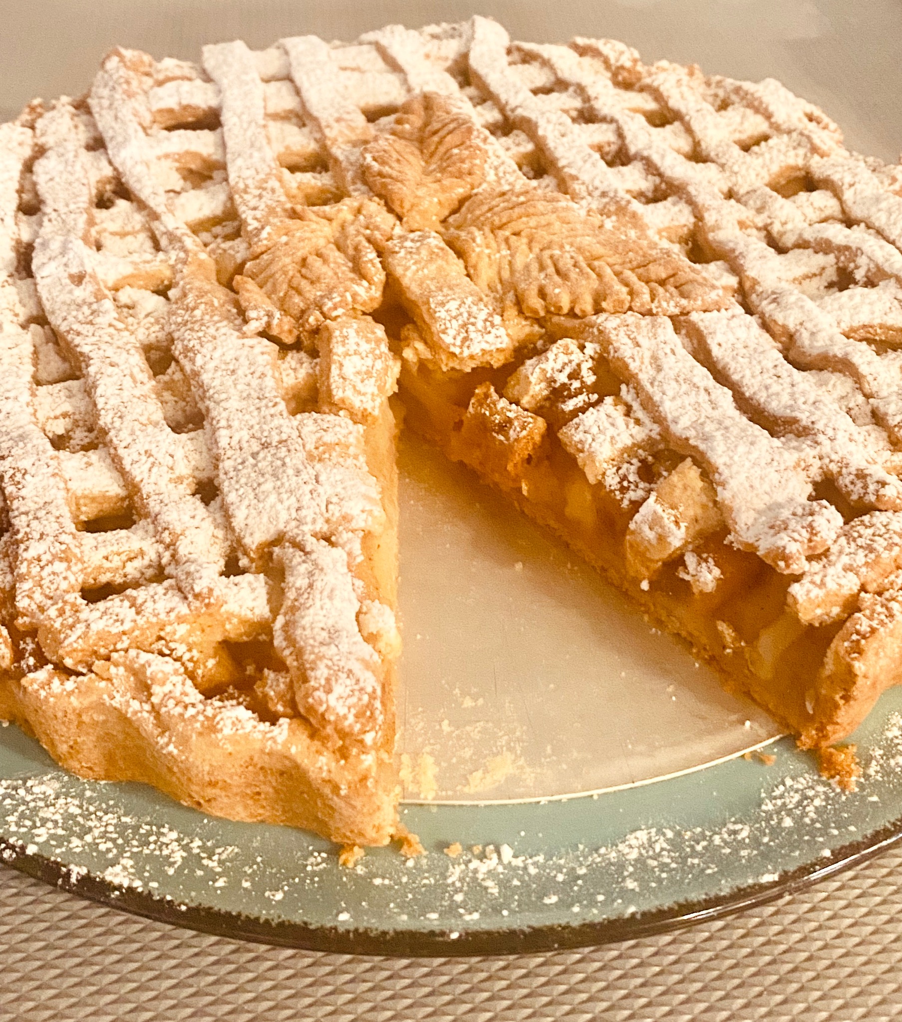 Jabłecznik z kratką posypany cukrem pudrem, wycięty kawałek, na błękitnym talerzu. Widoczne kawałki jabłek w nadzieniu. Całość w ciepłej tonacji.