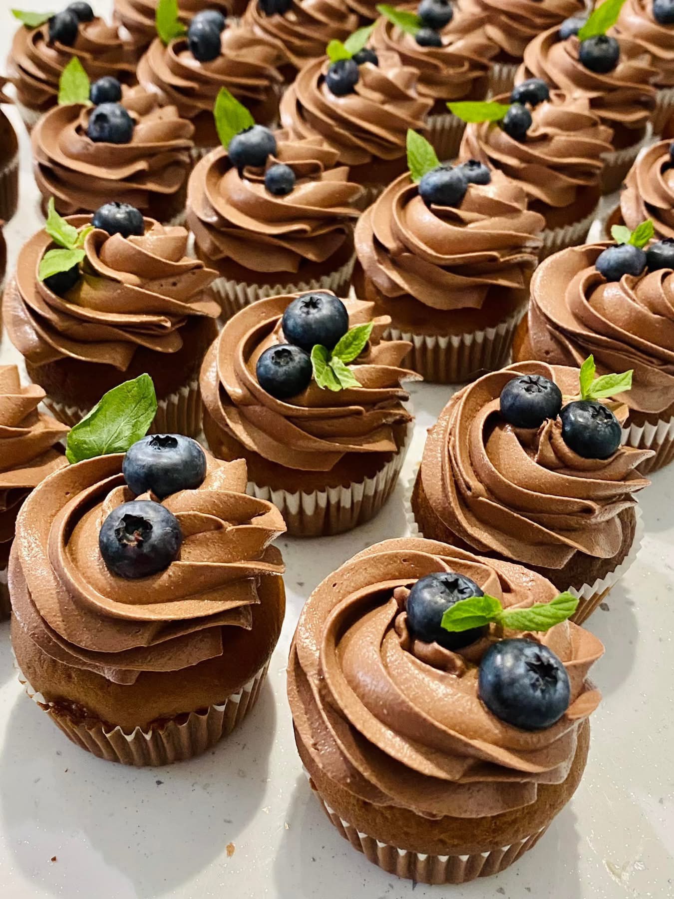 Apetyczne babeczki z kremem czekoladowym i borówkami, udekorowane listkami mięty, prezentowane przez cukiernię z Gdańska. Słodki poczęstunek.