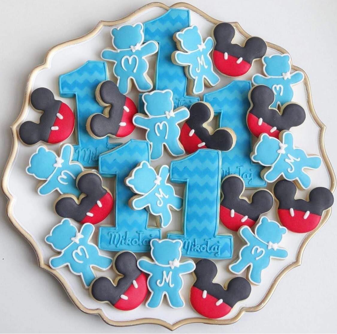 Dekoracyjne ciasteczka na roczek: błękitne cyfry '1', misie z literką 'M' i głowy Myszki Miki na białej paterze z delikatnym złotym obramowaniem.