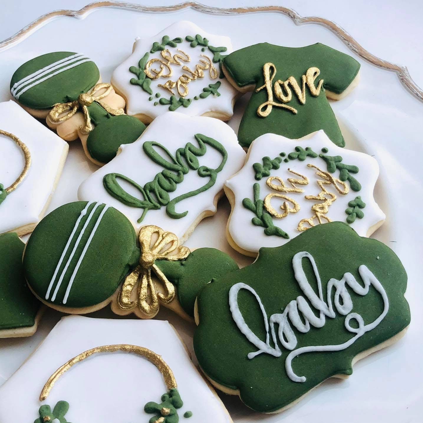 Dekoracyjne ciasteczka w odcieniach zieleni i złota na baby shower. Różne kształty: body, grzechotka, z napisem 'Baby' i 'Love'. Kremowe tło talerza.