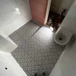 Jakub Kalata Investments - Ukończona podłoga w łazience z wzorzystymi płytkami, widok z góry na białą armaturę i fragment różowej ściany. W tle widoczne narzędzia i akcesoria.