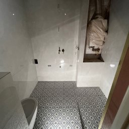 Jakub Kalata Investments - Biała łazienka z wzorzystą podłogą i częściowo wykończonymi ścianami. Widoczna instalacja wodna i kanalizacyjna, oraz fragment toalety po lewej stronie kadru.