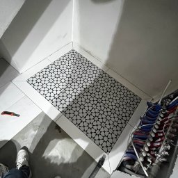 Jakub Kalata Investments - Ukończona podłoga w łazience z czarno-białą mozaiką w otoczeniu białych płytek. Widoczne rury instalacji wodnej i narzędzie glazurnicze.