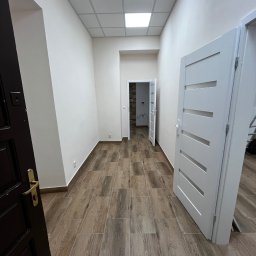 Jakub Kalata Investments - Wykończone wnętrze korytarza z drewnopodobną podłogą, białymi ścianami i widocznymi drzwiami. Widok w perspektywie, z otwartymi drzwiami w tle.