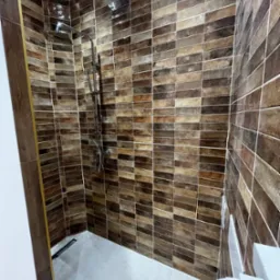 Nowoczesna kabina prysznicowa z brązowymi, prostokątnymi płytkami ułożonymi poziomo i odpływem liniowym. Całość utrzymana w ciepłej tonacji.