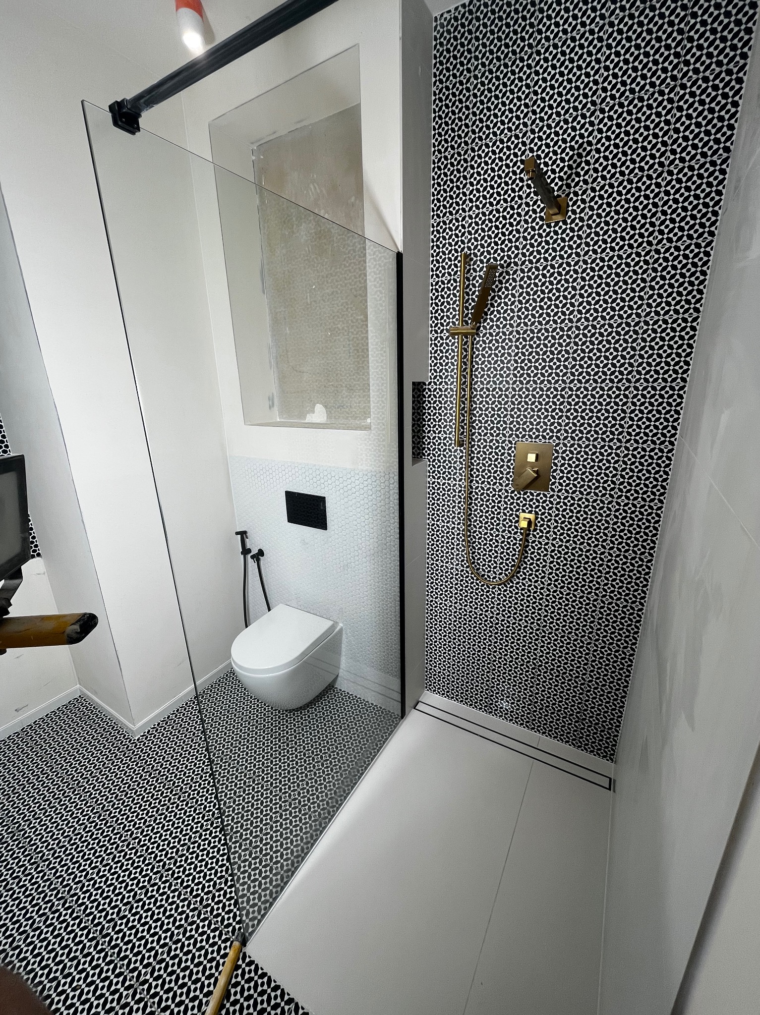 Nowoczesna łazienka po remoncie: czarno-biała mozaika na podłodze i ścianie prysznica, złota armatura, minimalistyczna toaleta podtynkowa. Kabina prysznicowa ze szklaną ścianką.