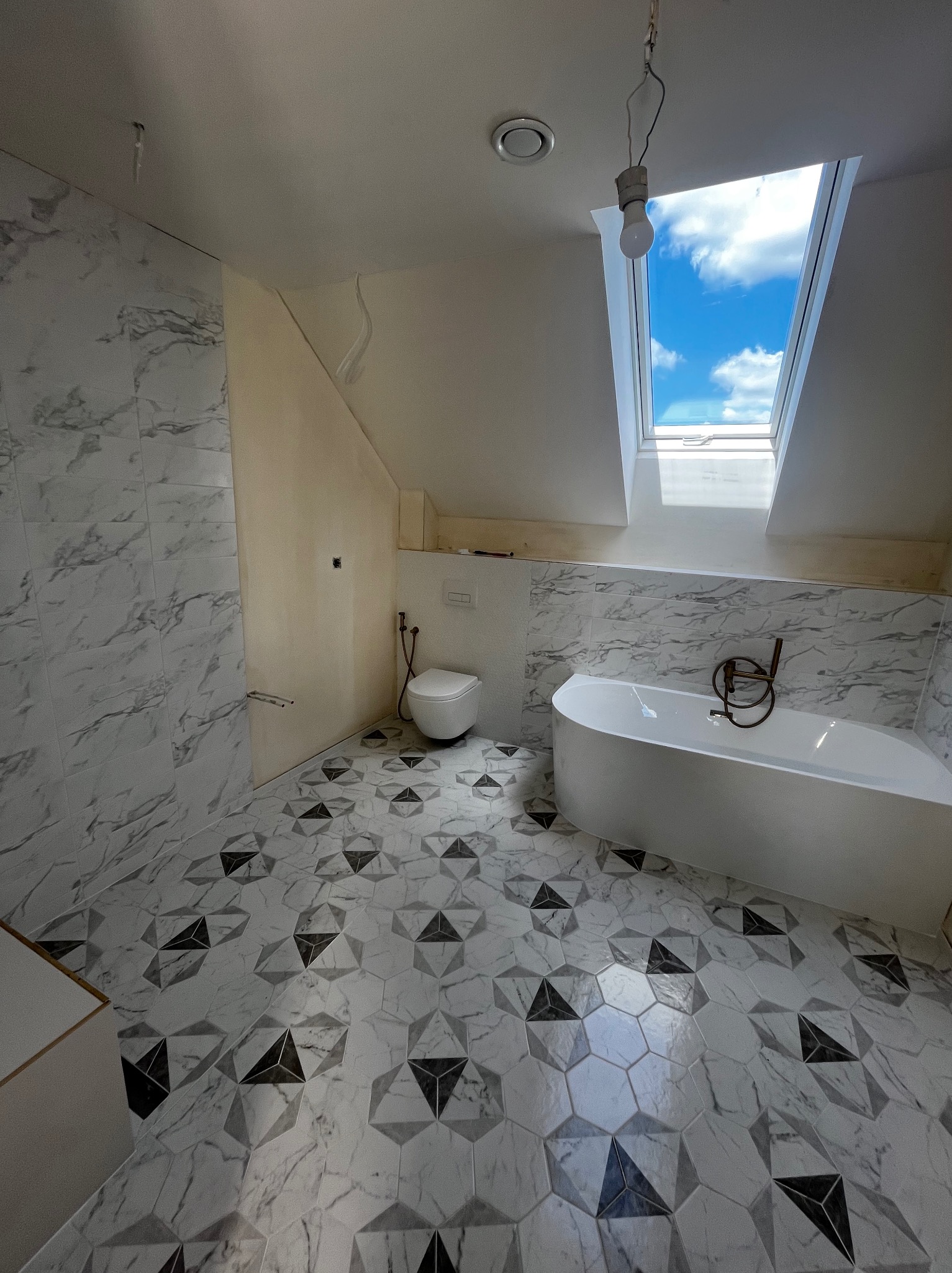 Ukończona łazienka na poddaszu z oknem dachowym. Marmurowe płytki ścienne i heksagonalna podłoga z geometrycznym wzorem. Biała wanna i toaleta.