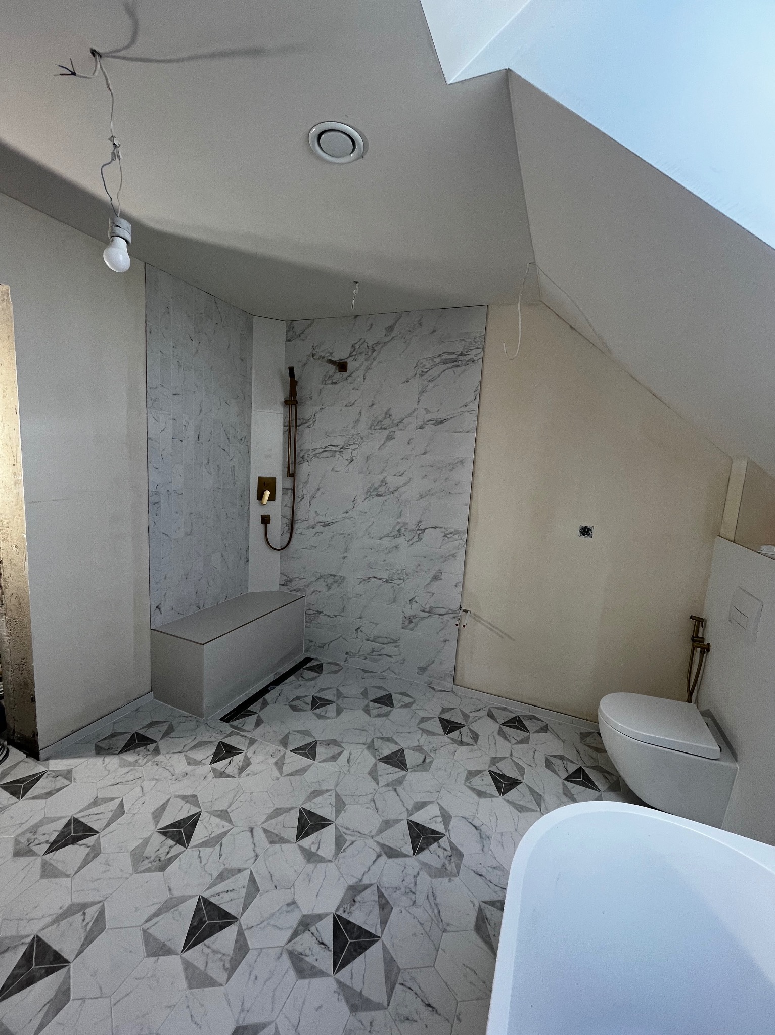 Łazienka na poddaszu z marmurowymi płytkami, mozaikową podłogą i złotą armaturą. Widoczna wanna, toaleta i prysznic. Oświetlenie sufitowe i wisząca żarówka.