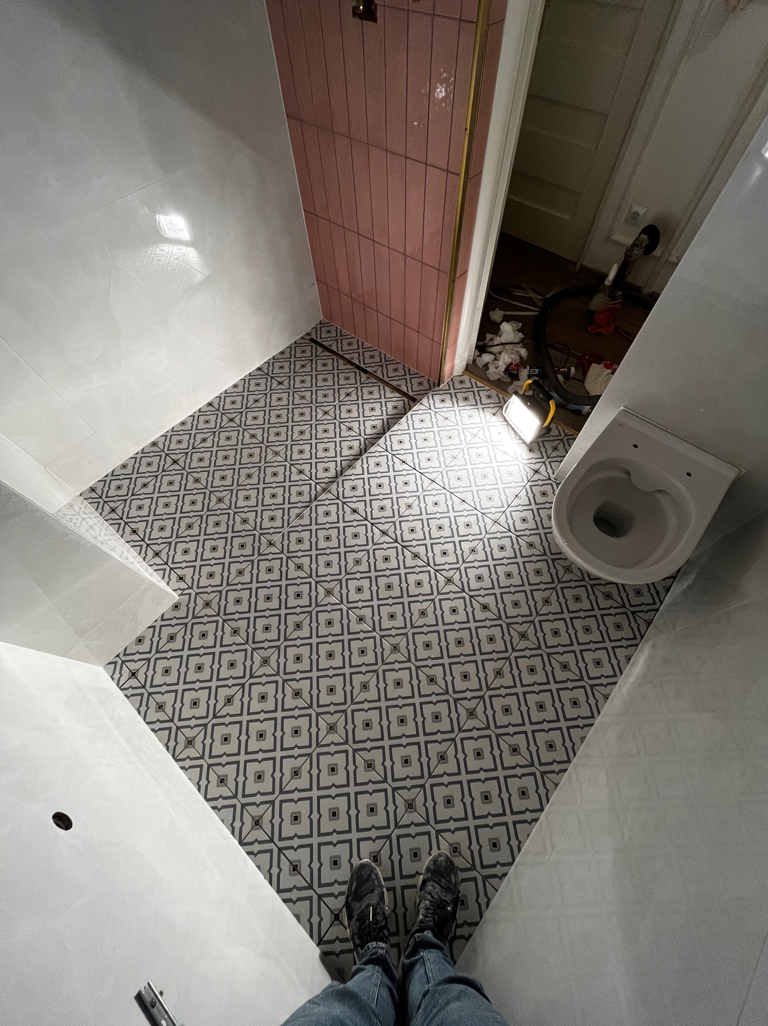 Ukończona podłoga w łazience z wzorzystymi płytkami, widok z góry na białą armaturę i fragment różowej ściany. W tle widoczne narzędzia i akcesoria.
