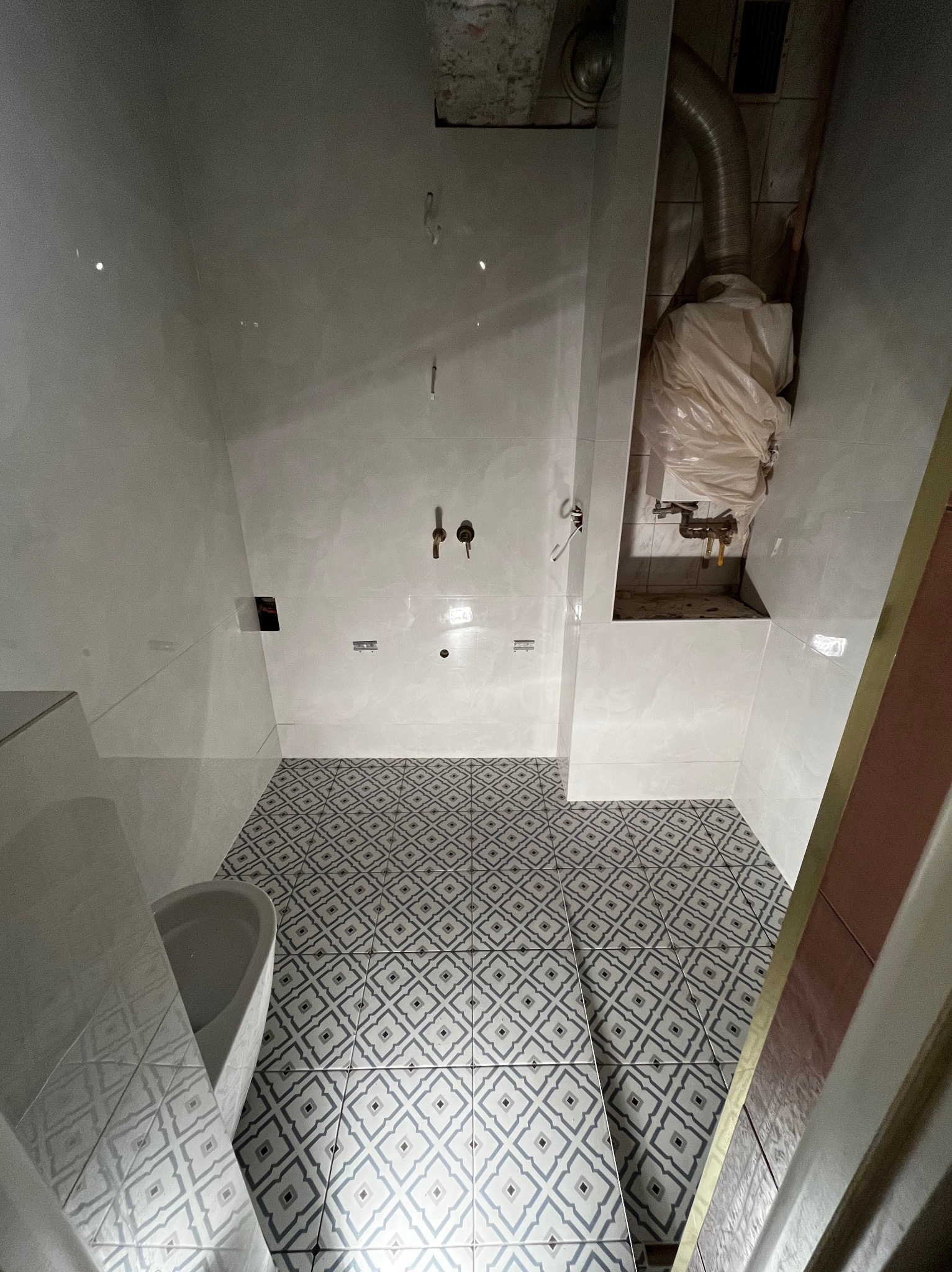 Biała łazienka z wzorzystą podłogą i częściowo wykończonymi ścianami. Widoczna instalacja wodna i kanalizacyjna, oraz fragment toalety po lewej stronie kadru.