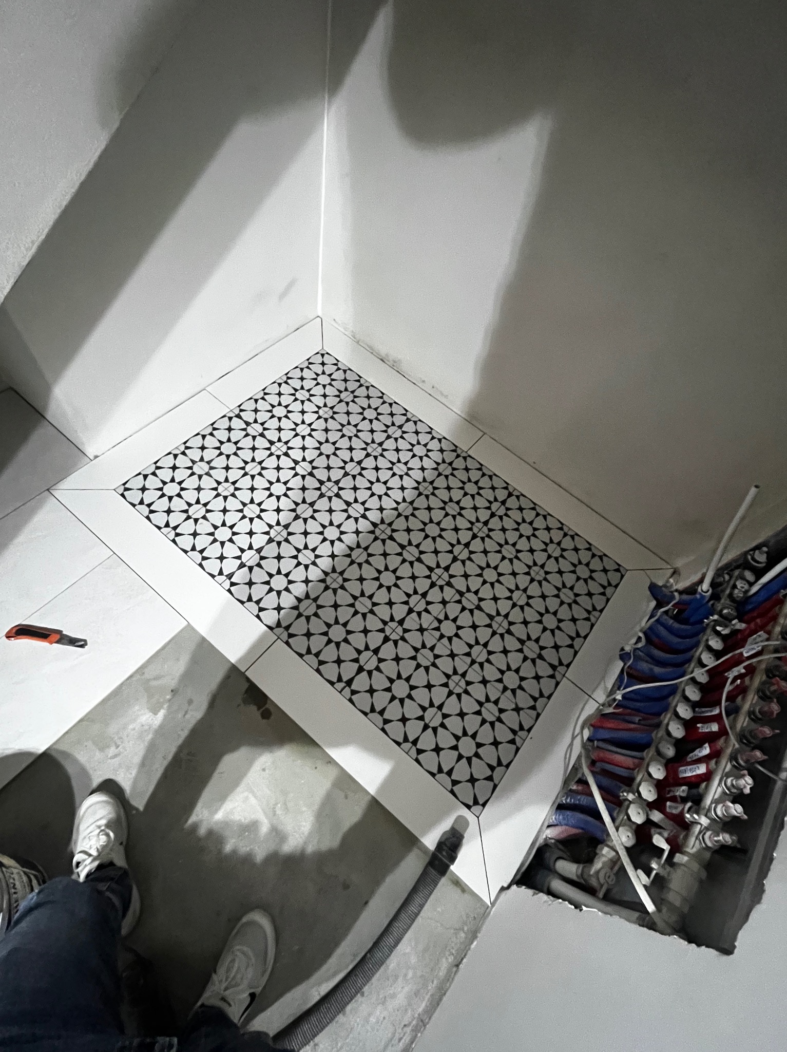 Ukończona podłoga w łazience z czarno-białą mozaiką w otoczeniu białych płytek. Widoczne rury instalacji wodnej i narzędzie glazurnicze.