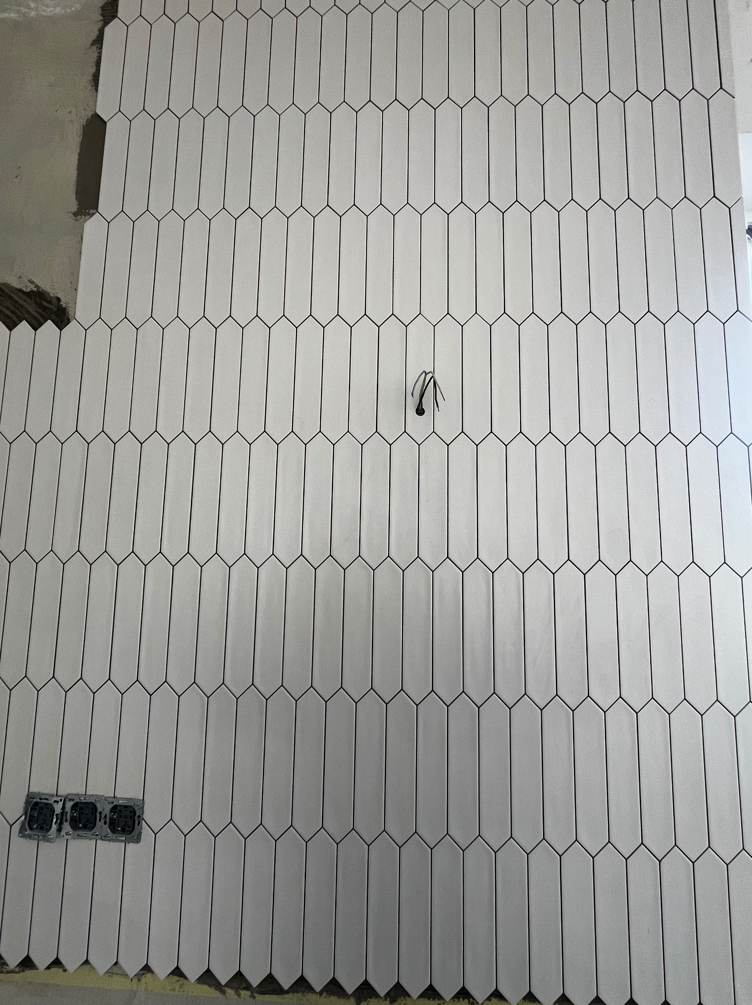 Ściana wyłożona białymi, podłużnymi płytkami w kształcie migdałów, z widocznymi puszkami elektrycznymi i fragmentem nieukończonej powierzchni z resztkami zaprawy.