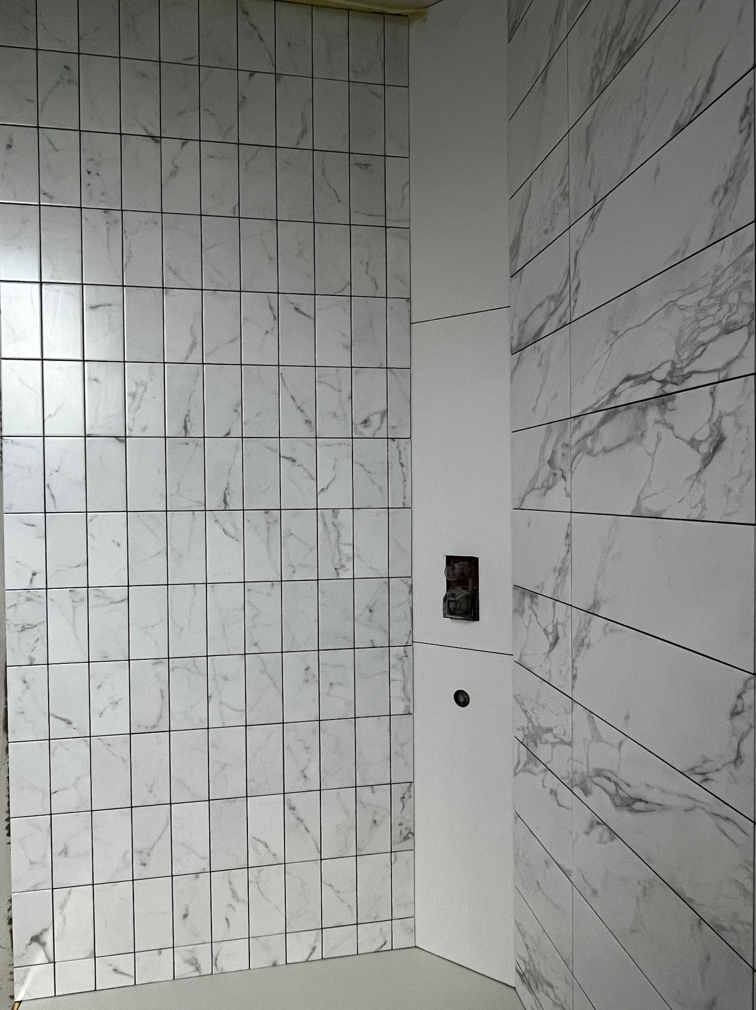 Ściana łazienki wyłożona białymi płytkami imitującymi marmur, ułożonymi w regularny wzór. Widoczne gniazdko elektryczne i otwór na rurę, co wskazuje na etap prac wykończeniowych.