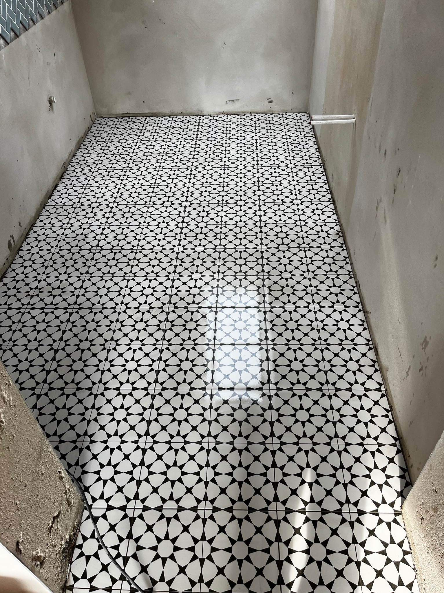 Ułożona podłoga z płytek w biało-czarny wzór geometryczny, widok z góry. Surowe ściany w trakcie wykończenia, widoczne instalacje.