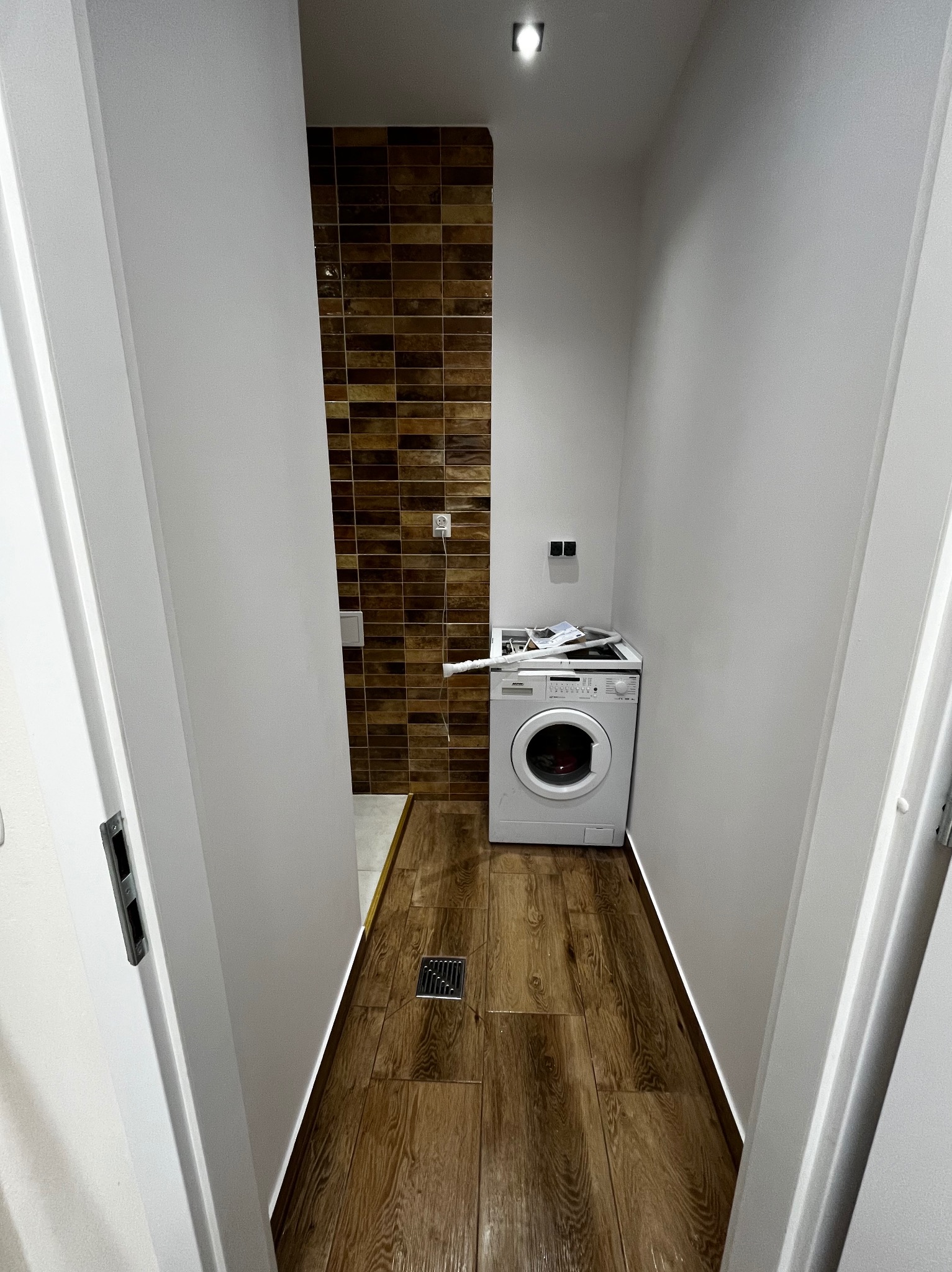 Wykończona łazienka z brązowymi płytkami na ścianie i drewnopodobną podłogą. Biała pralka w rogu. Widok zza otwartych białych drzwi.