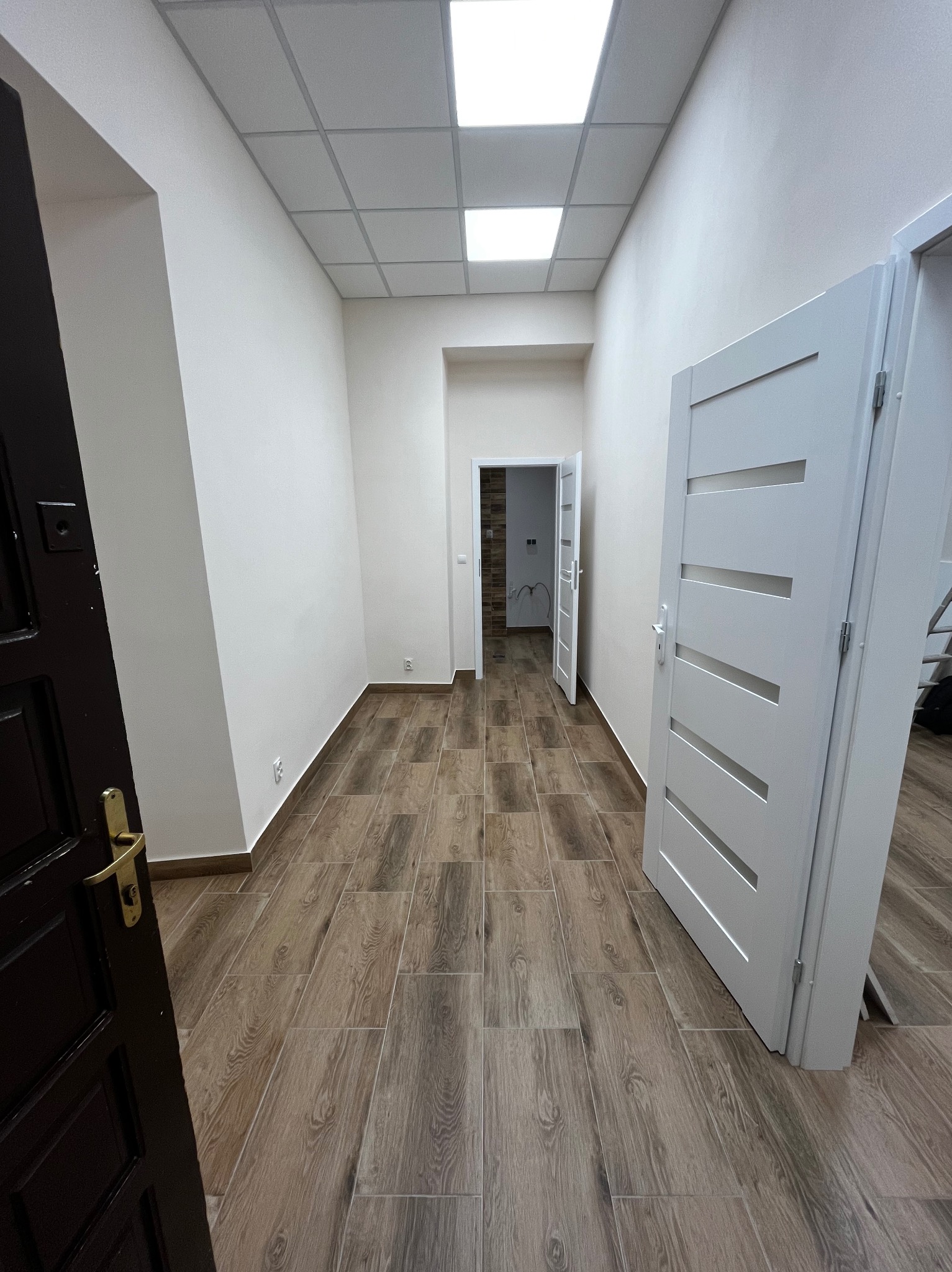 Wykończone wnętrze korytarza z drewnopodobną podłogą, białymi ścianami i widocznymi drzwiami. Widok w perspektywie, z otwartymi drzwiami w tle.