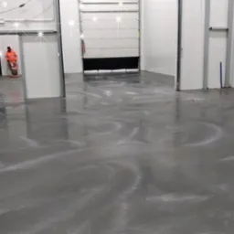 Gładka, szara posadzka z delikatnym wzorem w hali przemysłowej, widoczny pracownik w pomarańczowym kombinezonie w tle.