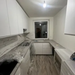 Jasna kuchnia po remoncie z białymi szafkami, marmurowym blatem i drewnopodobną podłogą. Część blatu zabezpieczona folią ochronną. Widok na okno.