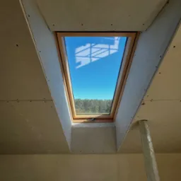 Montaż okna dachowego w skośnej ścianie z płyt kartonowo-gipsowych. Widok z dołu na błękitne niebo i drzewa. Częściowo widoczna podpora.