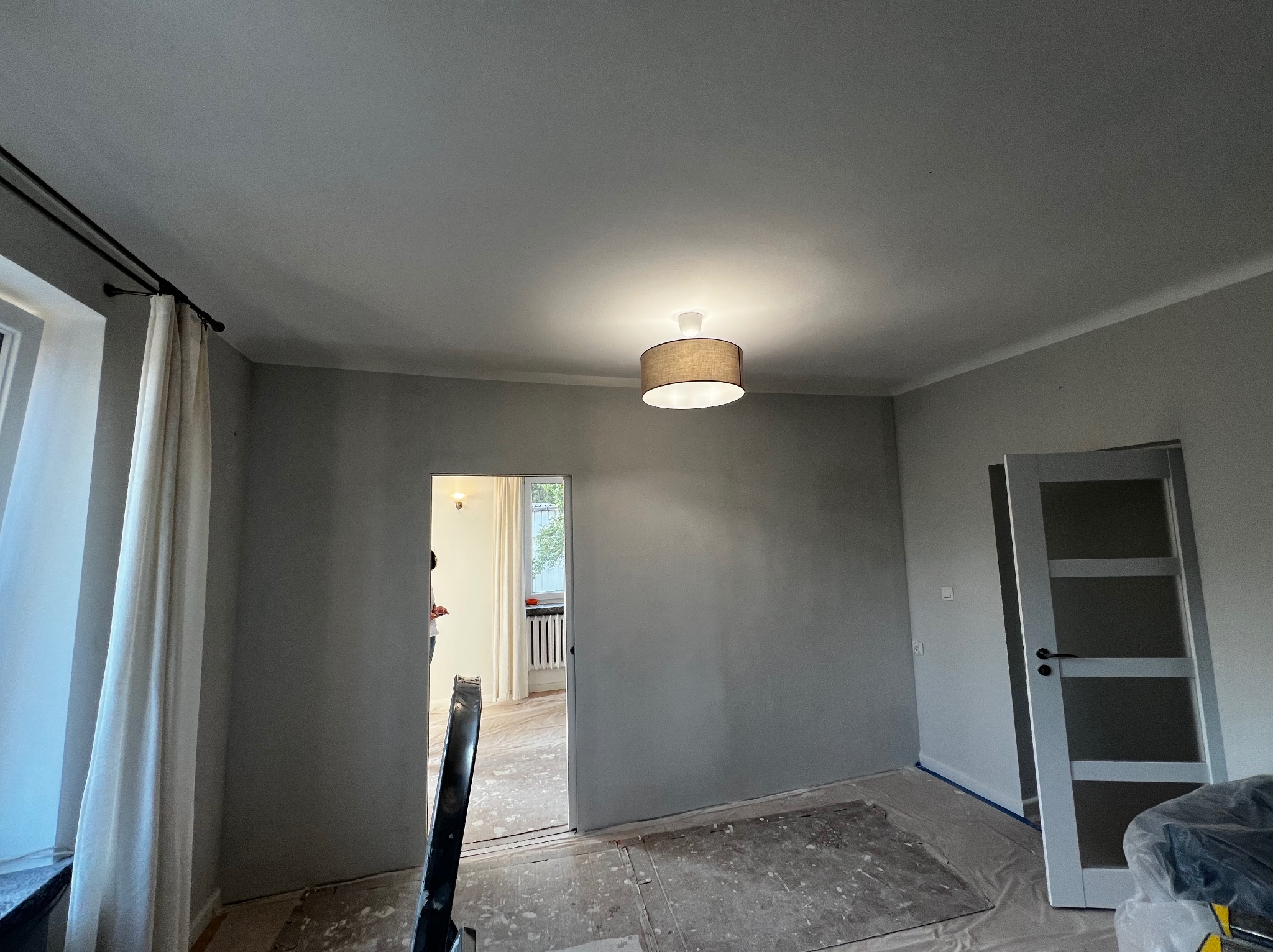 Wnętrze pokoju po malowaniu ścian na szaro, widoczne drzwi z przeszkleniami i lampa sufitowa. Podłoga zabezpieczona folią malarską. Widok na sąsiednie pomieszczenie.