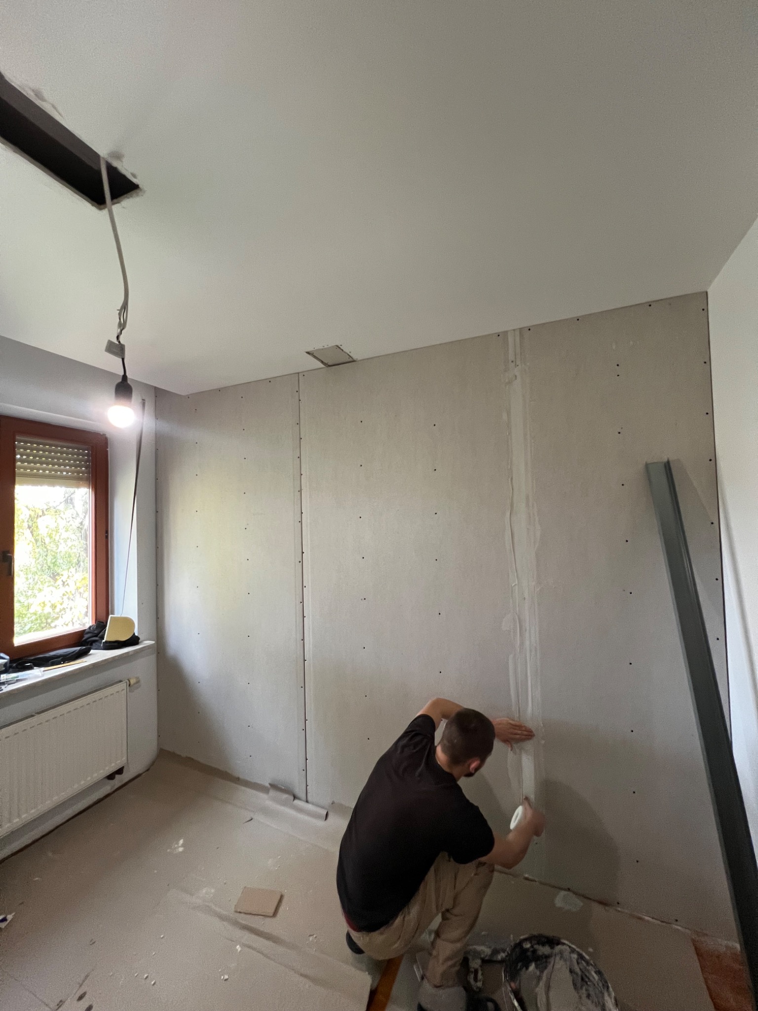 Prace wykończeniowe w pokoju: szpachlowanie łączeń płyt karton-gips na ścianie. Widoczne narzędzia, okno z żaluzjami i grzejnik. W tle surowy sufit z wystającymi kablami.