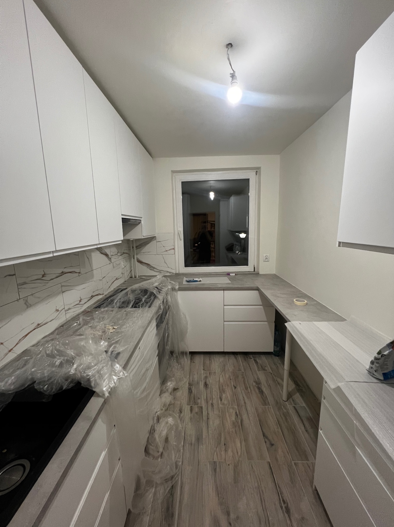 Jasna kuchnia po remoncie z białymi szafkami, marmurowym blatem i drewnopodobną podłogą. Część blatu zabezpieczona folią ochronną. Widok na okno.