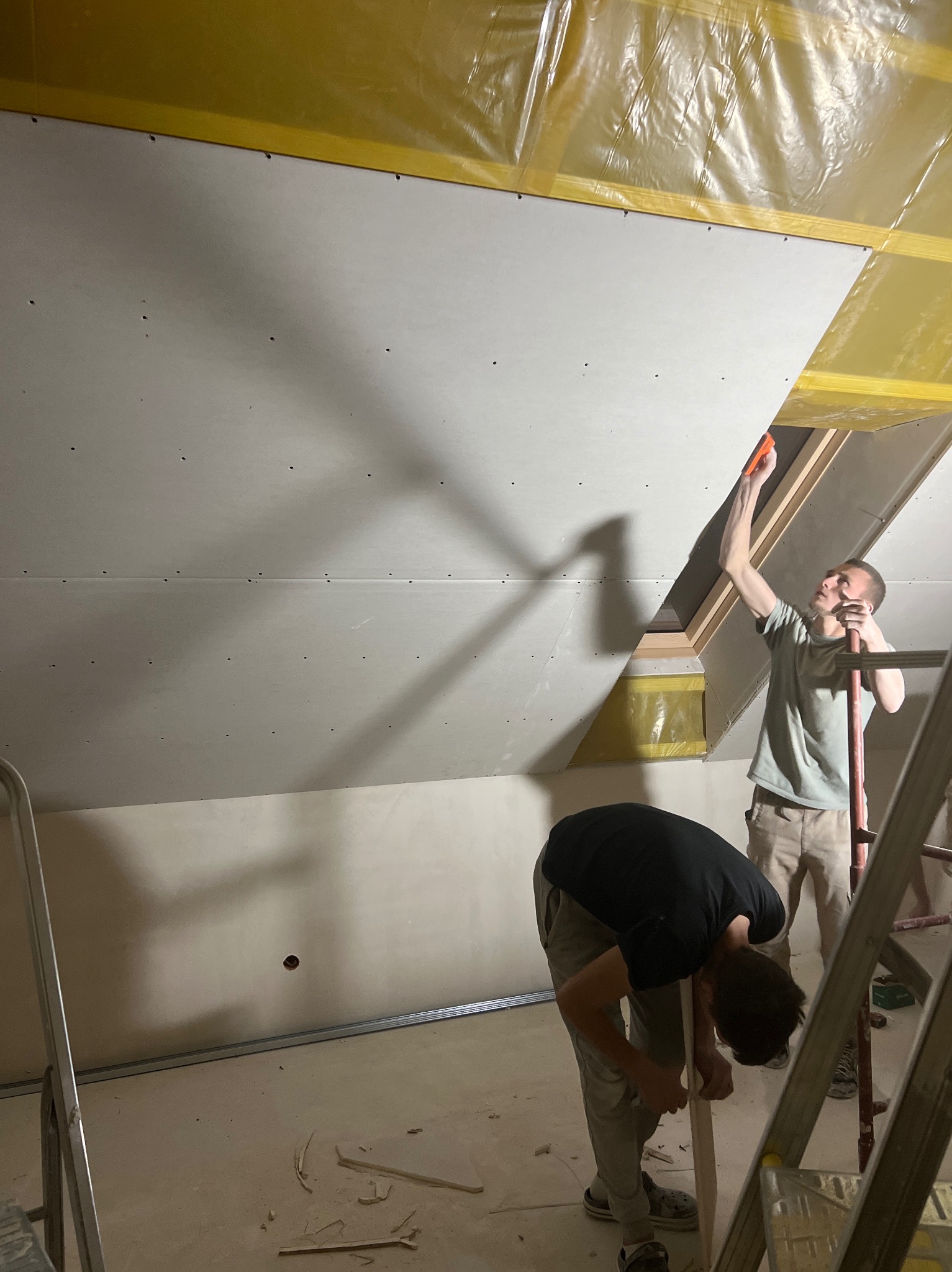 Montaż płyt gipsowo-kartonowych na poddaszu z oknem dachowym. Dwóch mężczyzn przy pracy, widoczne rusztowanie i narzędzia. Żółta folia izolacyjna na suficie.