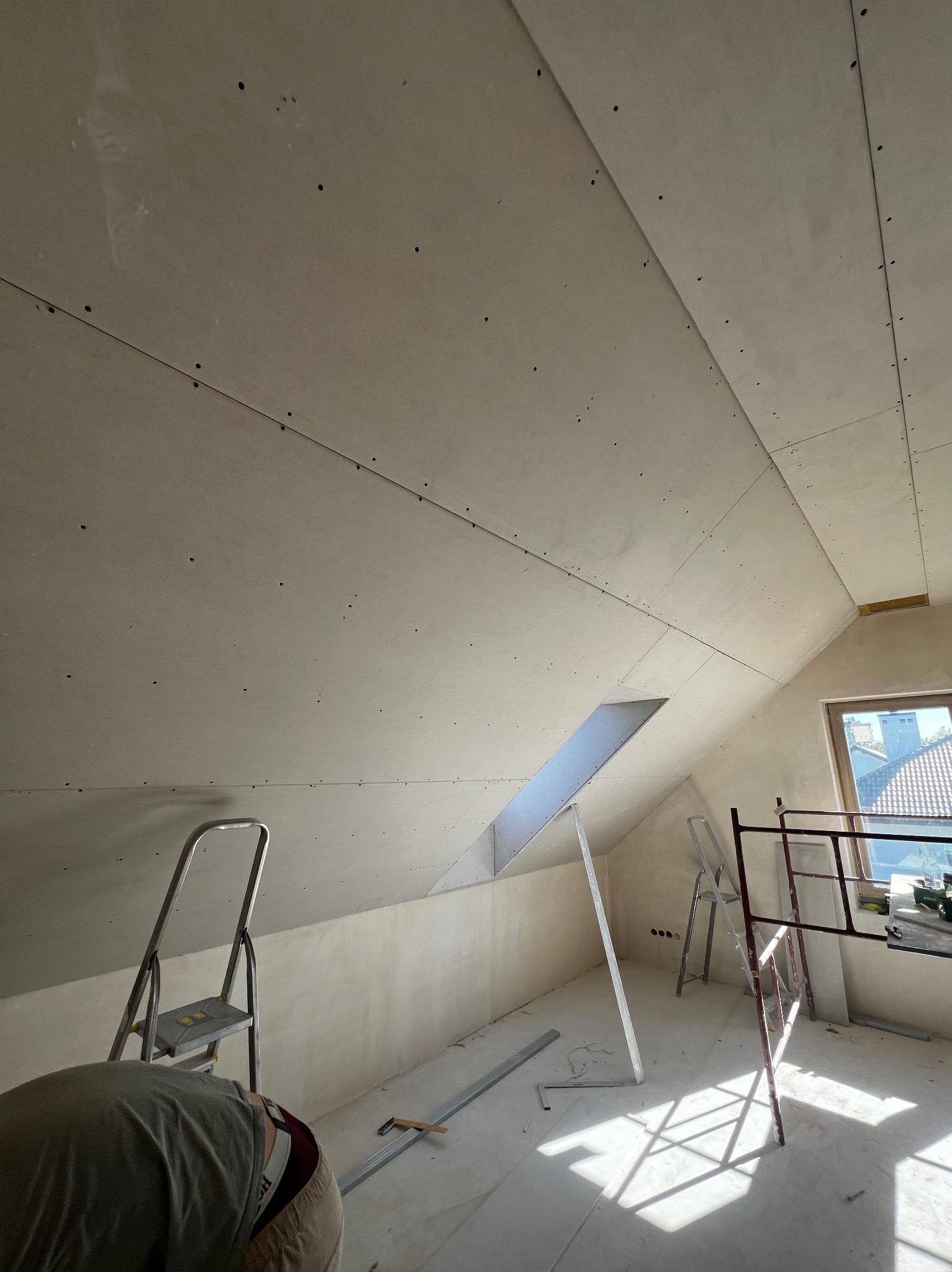 Poddasze w trakcie wykańczania płytami gipsowo-kartonowymi. Widoczne rusztowania, drabina i częściowo osoba wykonująca prace. Skos dachu z oknem.