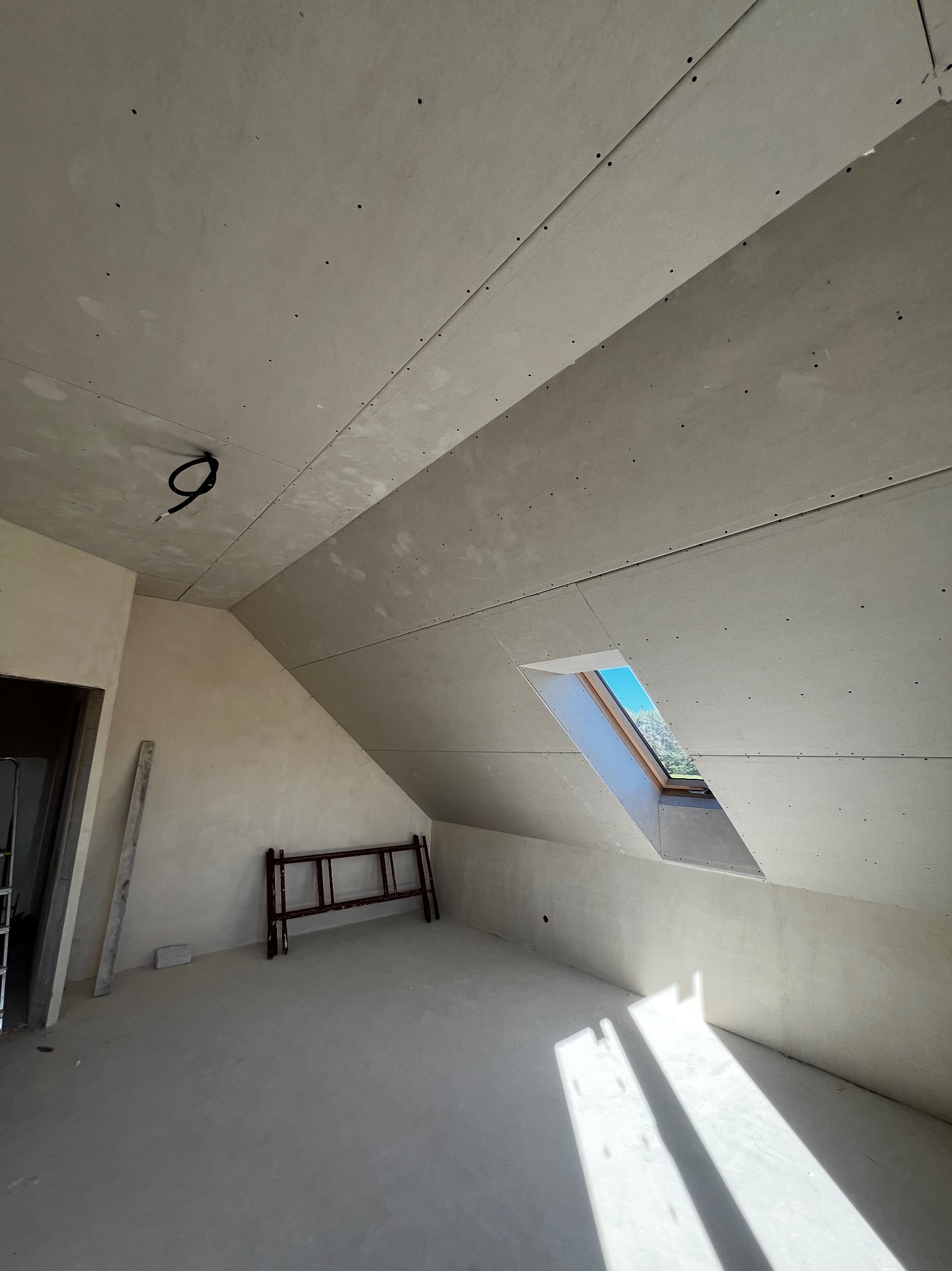 Poddasze z oknem dachowym w trakcie wykańczania płytami gipsowo-kartonowymi. Widoczne łączenia płyt, przewody elektryczne i drabina. Wpadające światło słoneczne.