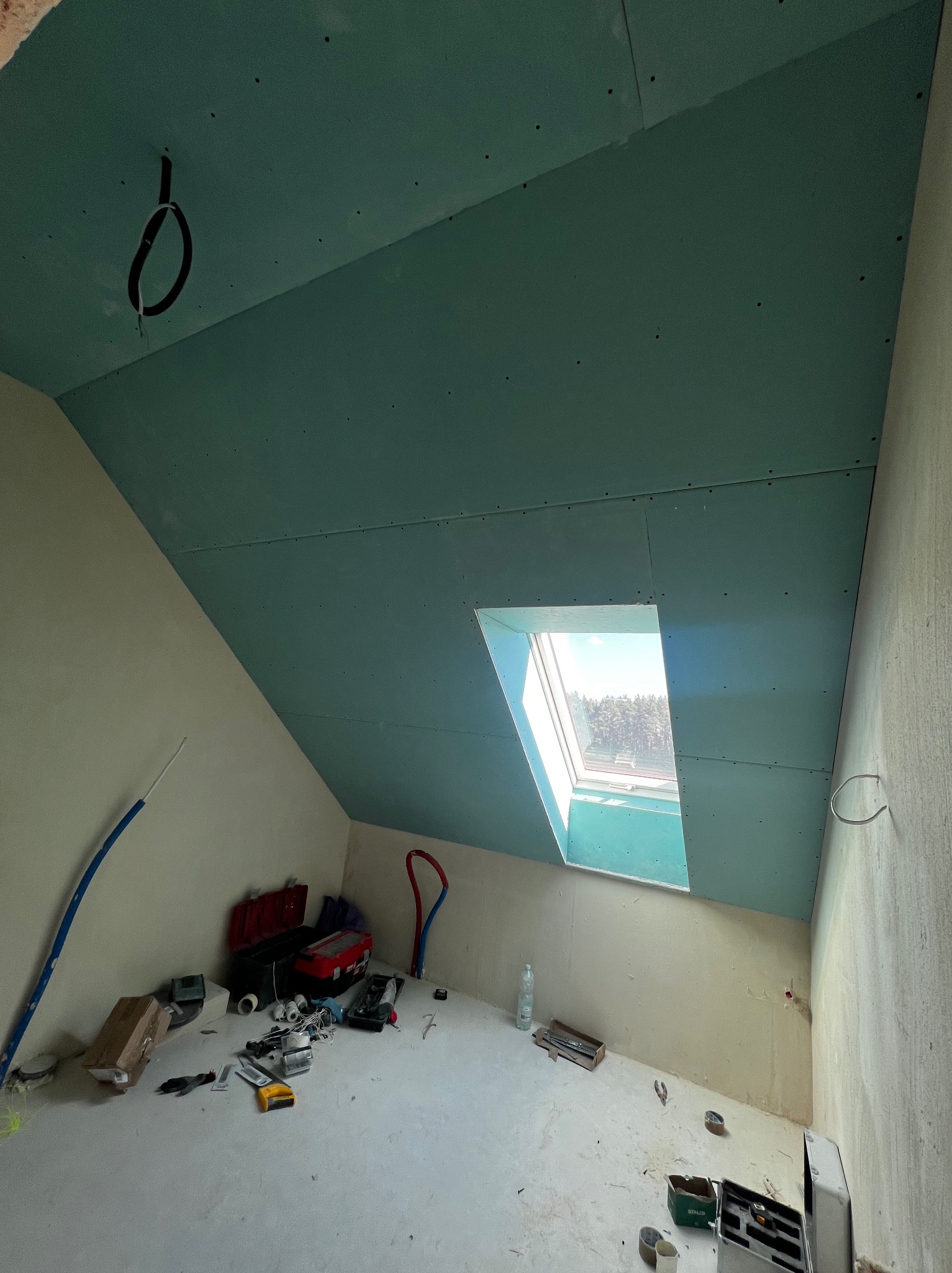 Poddasze z oknem dachowym po montażu zielonych płyt gipsowo-kartonowych. Widoczne narzędzia i akcesoria budowlane. Kabel zwisający z sufitu. Ściany przygotowane do dalszych prac.
