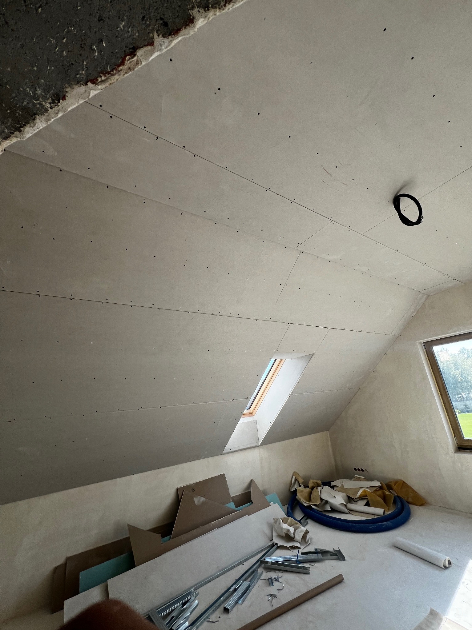 Poddasze z oknem dachowym po montażu płyt kartonowo-gipsowych na skosach i suficie. Widoczne łączenia płyt i otwór na oświetlenie. Część materiałów budowlanych na podłodze.