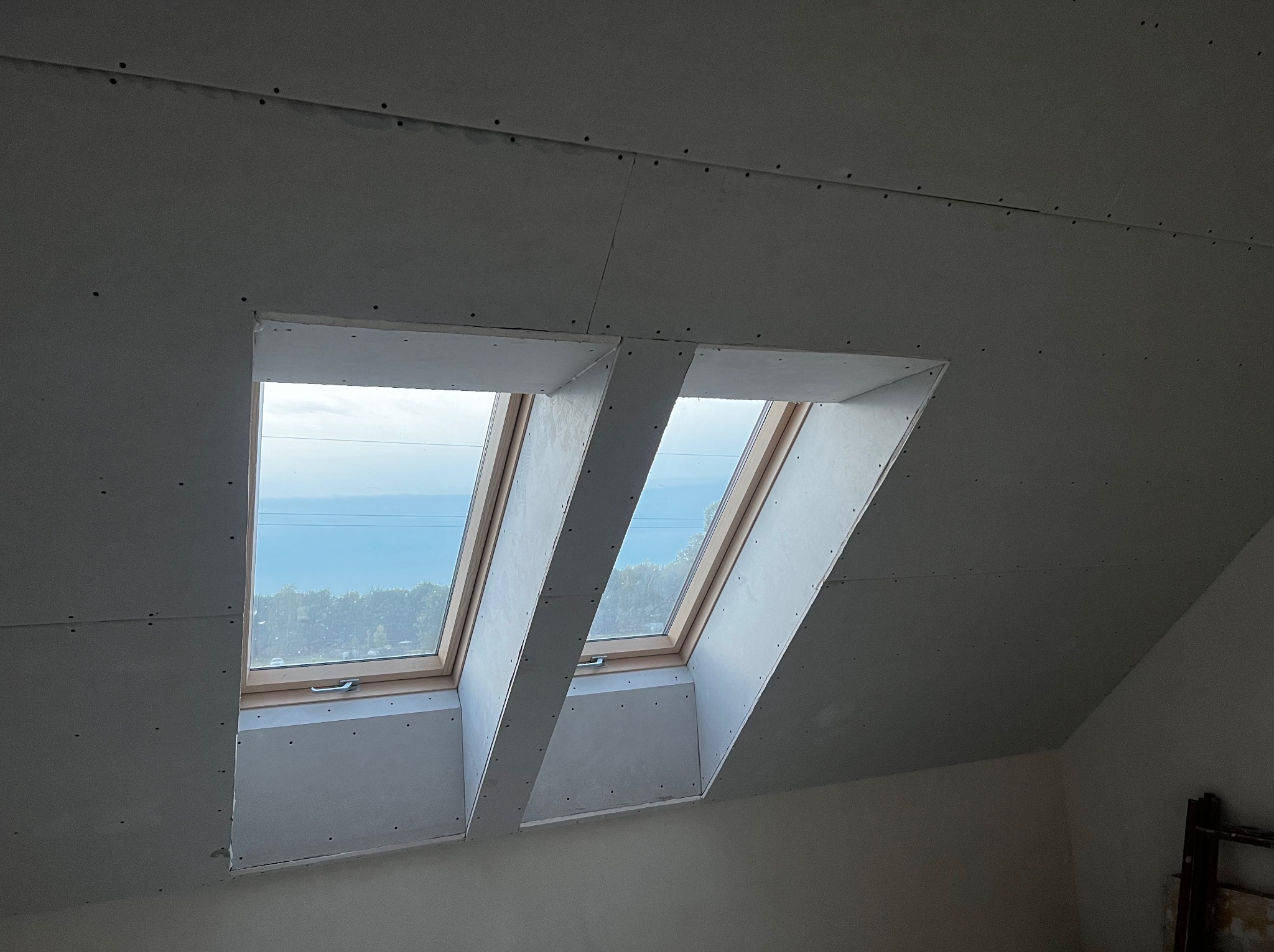 Montaż płyt karton gips na poddaszu wokół okien dachowych. Widoczne łączenia płyt i wkręty. Przez okna widać zieleń i niebo.
