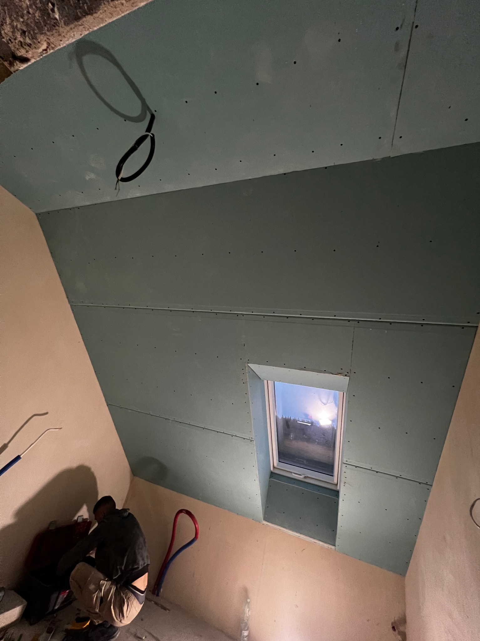 Montaż płyty G-K na poddaszu z oknem dachowym. Widoczne łączenia płyt i instalacja elektryczna. Fachowiec w trakcie pracy, narzędzia w tle. Ujęcie z dołu.