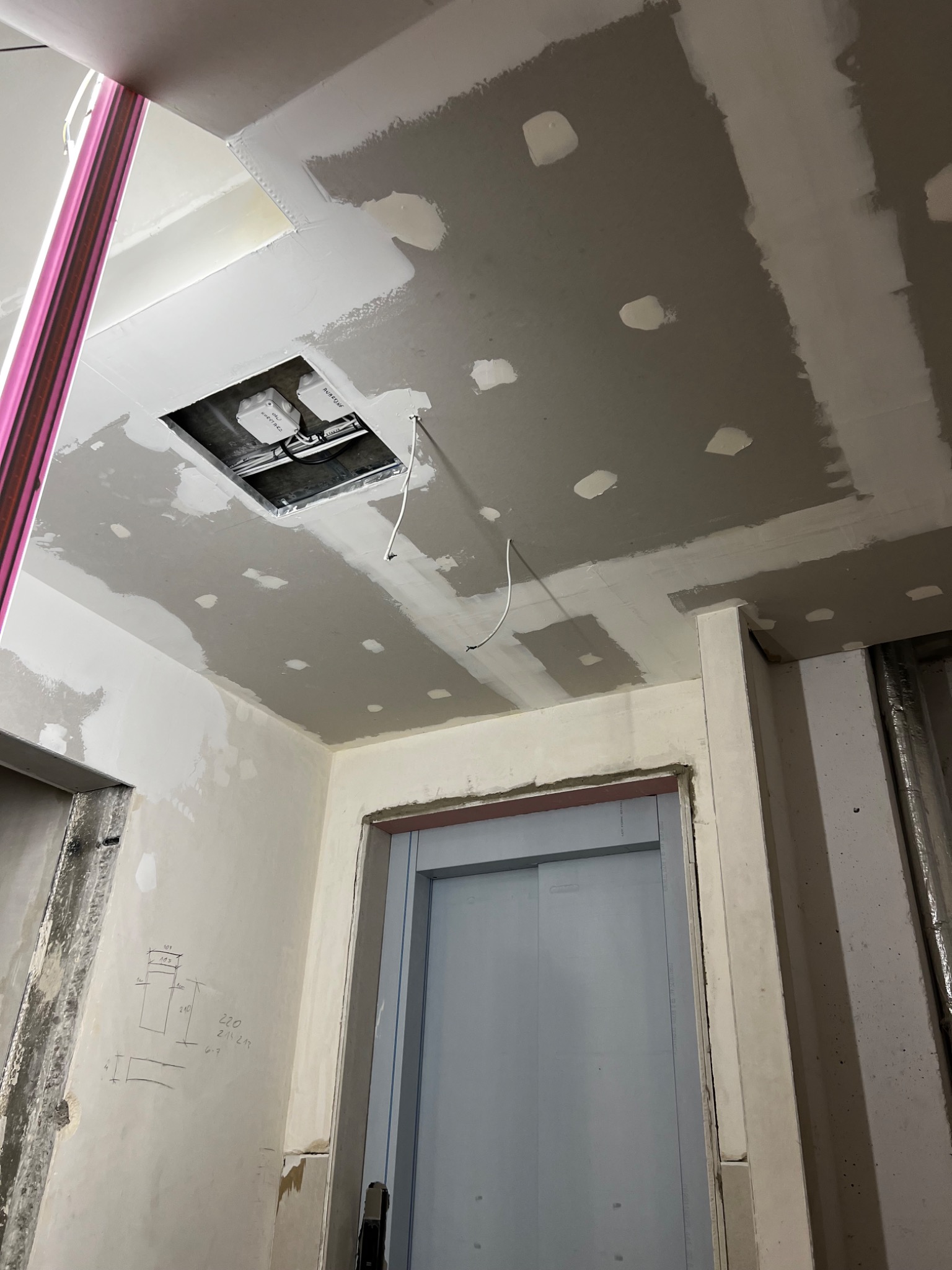 Sufit z płyt kartonowo-gipsowych w trakcie szpachlowania, widoczne łączenia i otwór rewizyjny z instalacją. Ściany w stanie surowym z odręcznymi notatkami.