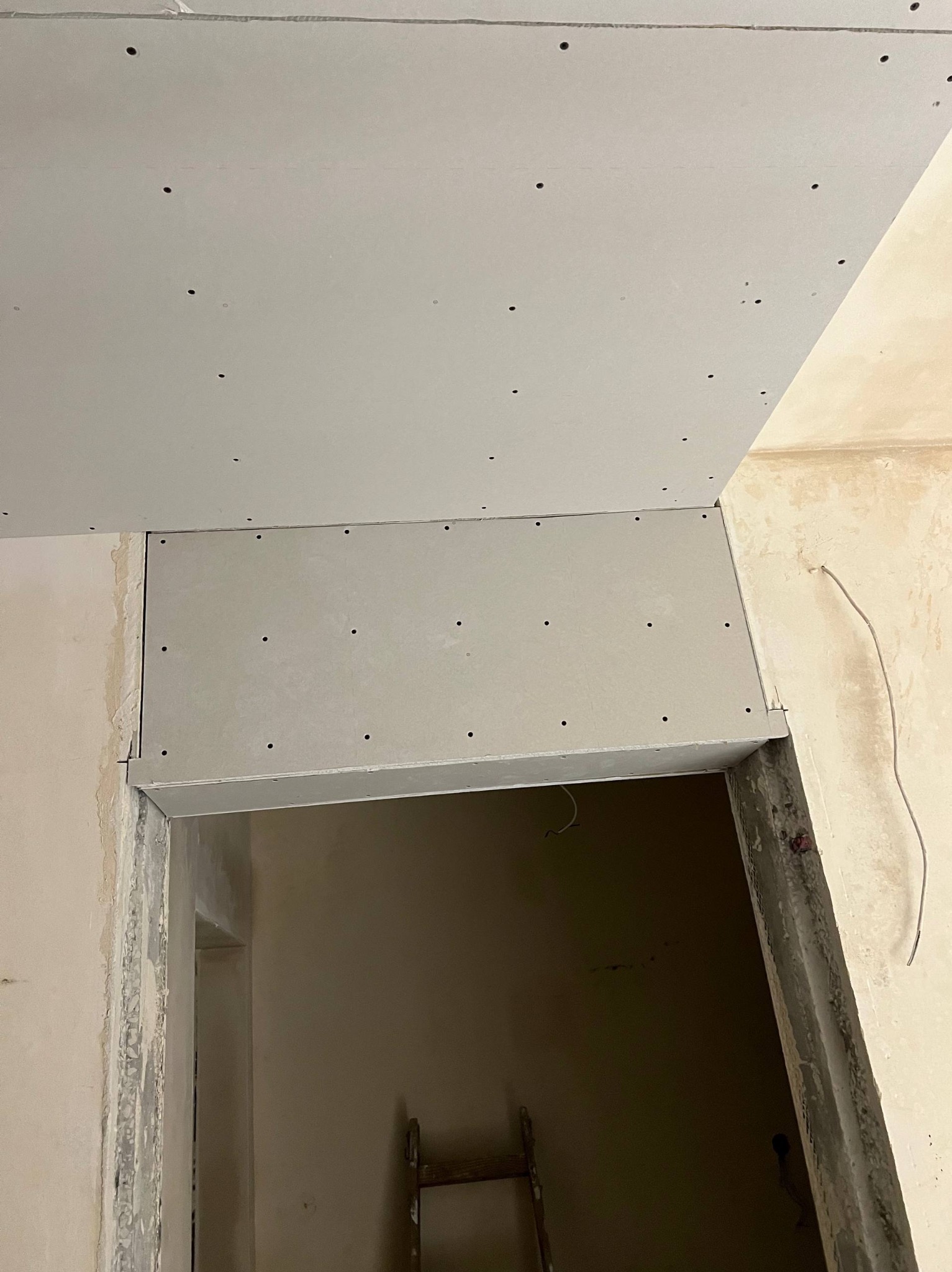 Montaż płyty gipsowo-kartonowej nad futryną drzwiową. Widoczne otwory na wkręty, surowe ściany i fragment drabiny w głębi pomieszczenia. Ujęcie z dołu.