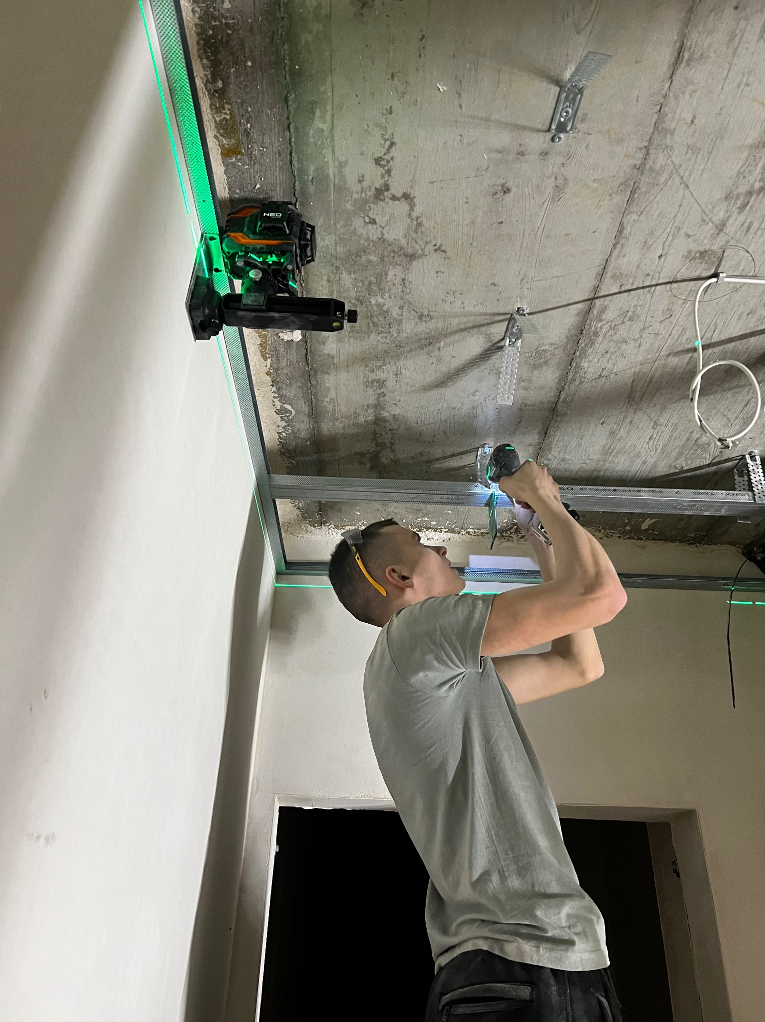 Pracownik montuje metalowy stelaż sufitu podwieszanego, używając wkrętarki i poziomicy laserowej z zielonym światłem. Widoczne profile i uchwyty mocujące do sufitu.