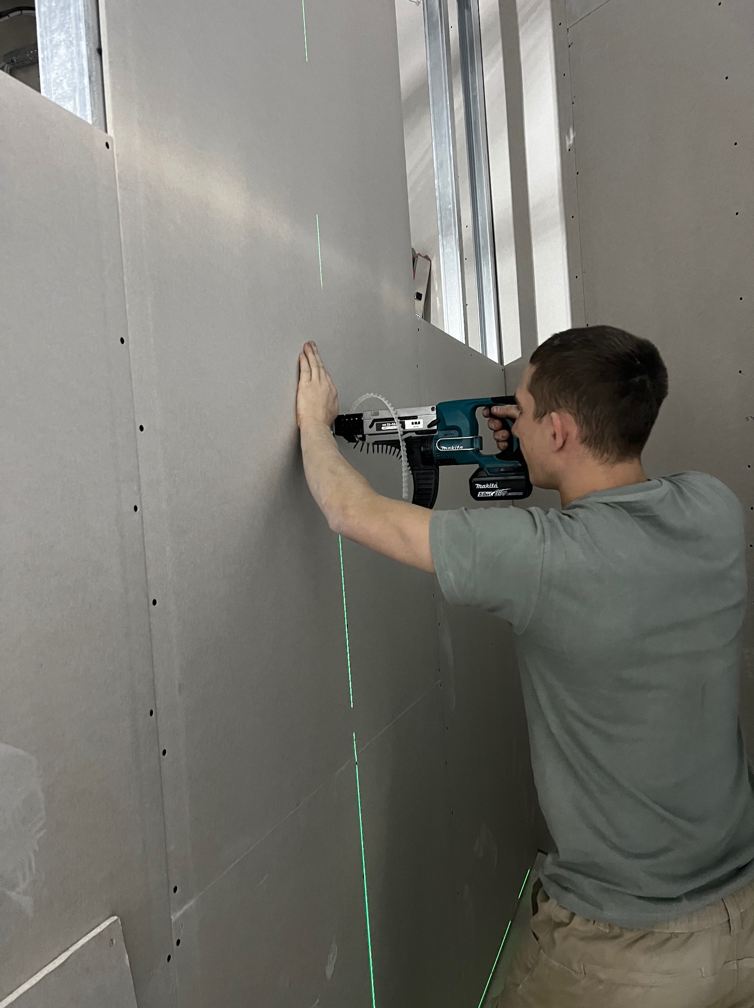 Pracownik montuje płytę karton gips na metalowym stelażu, używając wkrętarki akumulatorowej Makita i lasera krzyżowego do precyzyjnego ustawienia. Widoczne śruby mocujące.