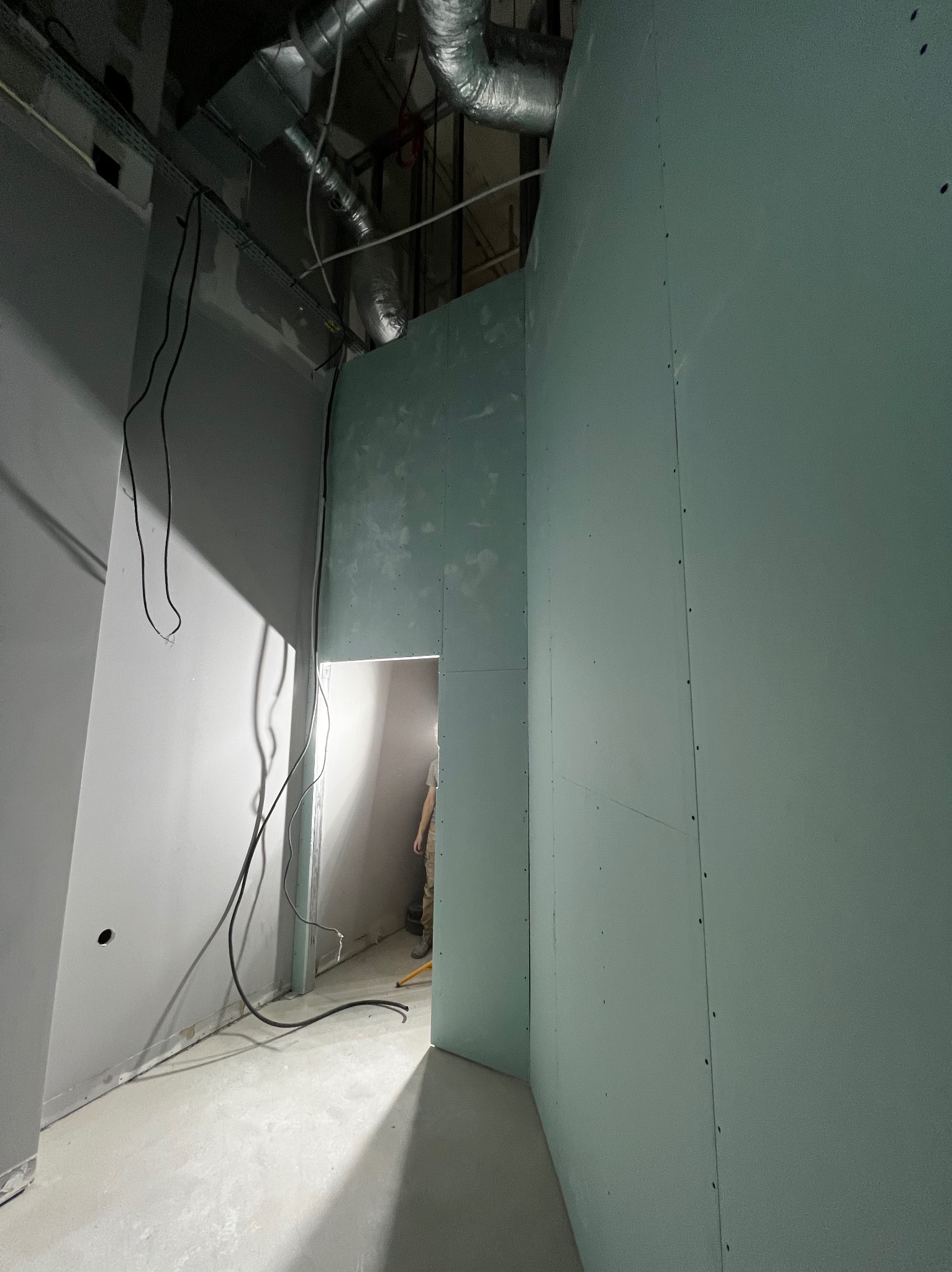 Wnętrze w trakcie prac wykończeniowych: ściany z płyt karton-gips, widoczne przewody i fragment instalacji wentylacyjnej pod sufitem. W tle sylwetka osoby.