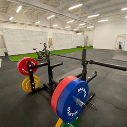 Marcin Braniewski - Profesjonalna siłownia: stojak ze sztangą obciążony kolorowymi talerzami Manto, w tle sprzęt do ćwiczeń, sztuczna trawa i drabina.