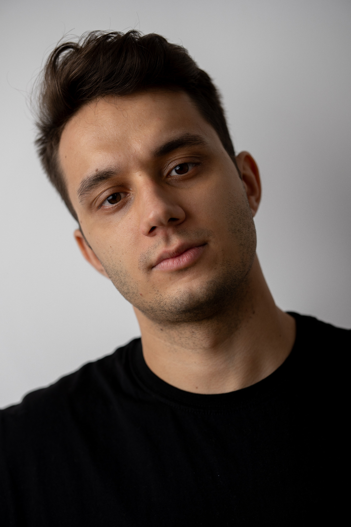 Młody mężczyzna w czarnej koszulce, z krótką brodą i zaczesanymi włosami, patrzy w obiektyw na jasnym tle. Portret w neutralnym oświetleniu.