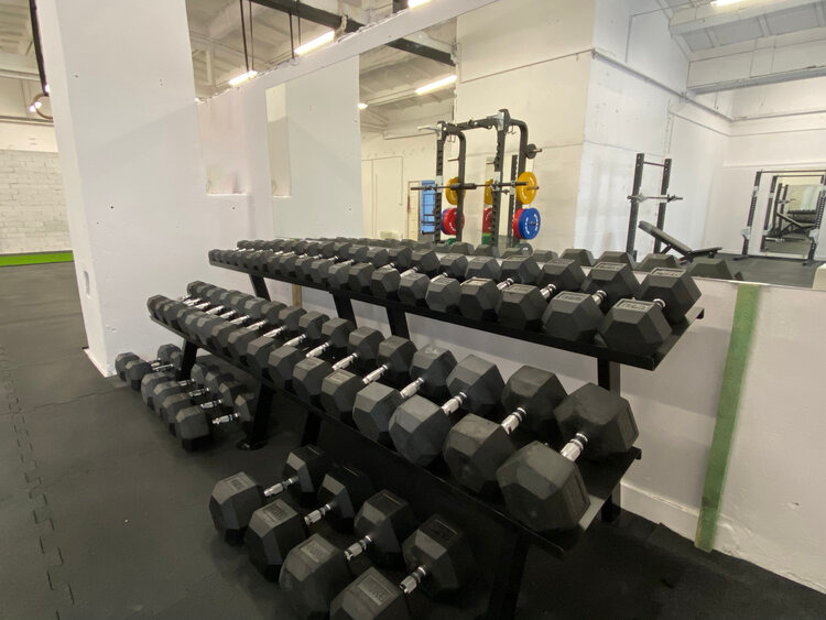Rzędy czarnych hantli na stojakach w jasnej siłowni. W tle lustro odbija sprzęt do ćwiczeń siłowych i obciążenia. Podłoga z czarnej gumy.