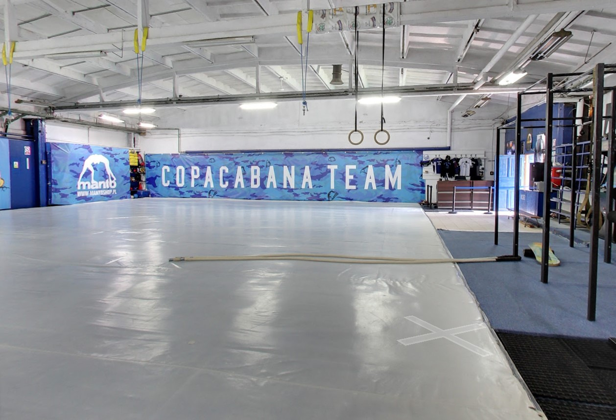 Jasna sala treningowa z szarą matą, wiszącymi obręczami gimnastycznymi i napisem 'Copacabana Team' na ścianie. Widoczne drabinki i sprzęt do ćwiczeń.