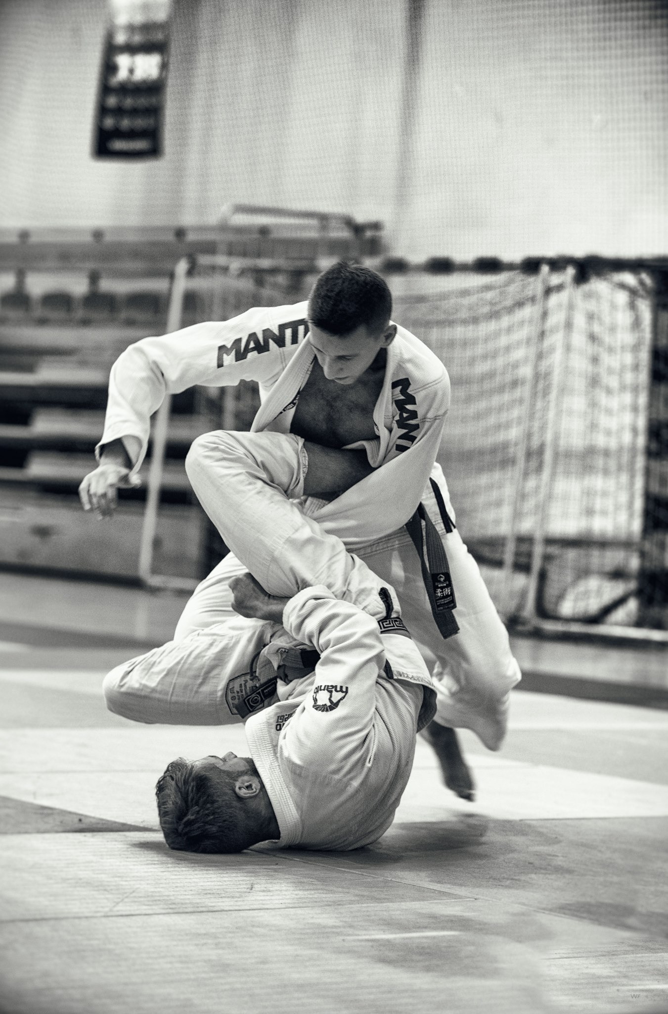 Czarno-białe zdjęcie dwóch mężczyzn w kimonach, podczas walki na macie. Jeden zawodnik unosi drugiego, wykonując dynamiczne poddanie. W tle siatka i elementy wyposażenia sali.