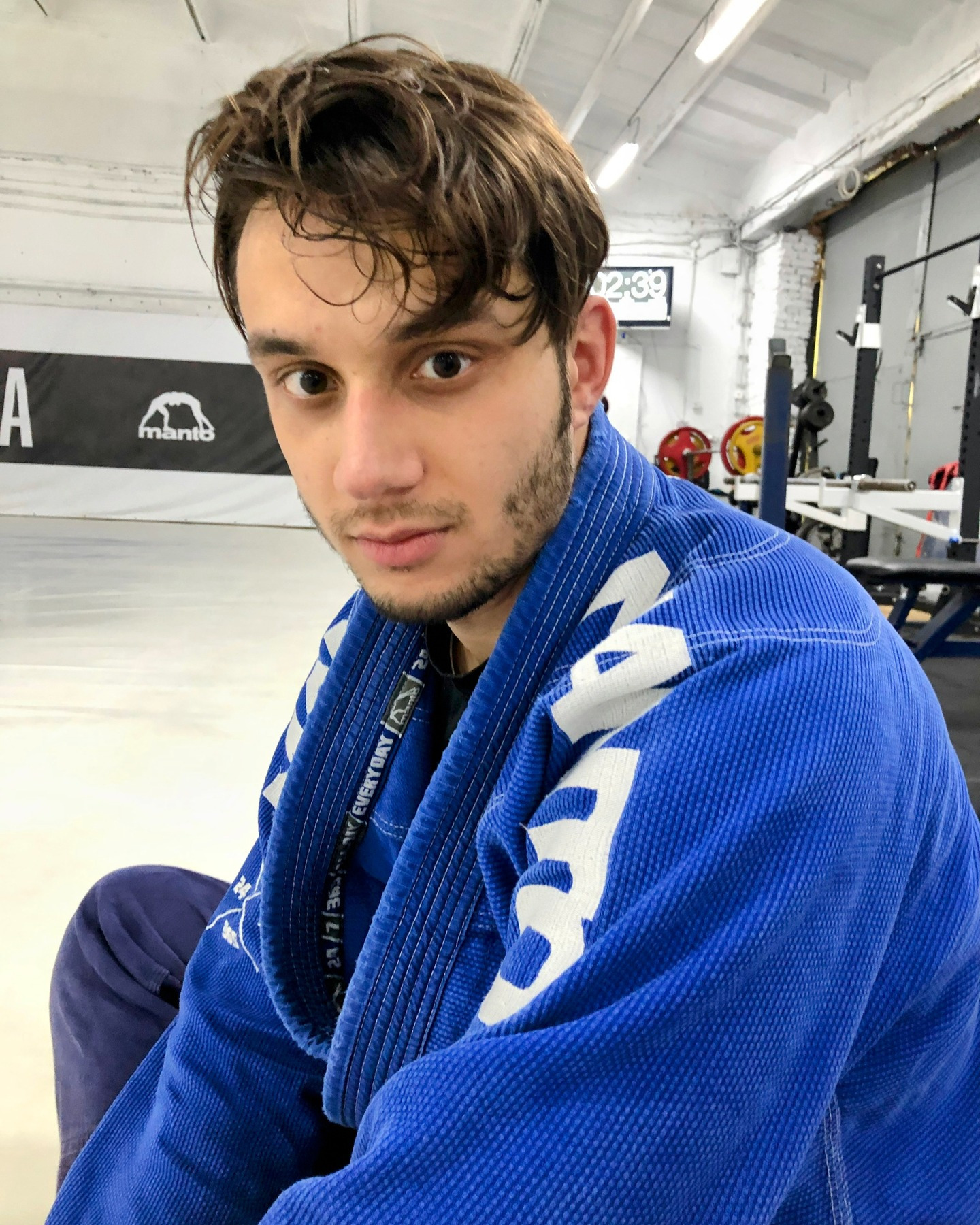 Młody mężczyzna w niebieskim kimonie do sportów walki, siedzący na sali treningowej z wyposażeniem siłowni w tle. Widoczny zegar i logo Manto.